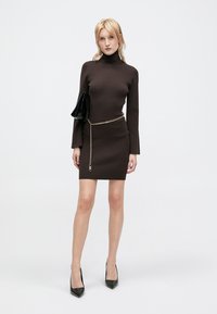 Brun ribbet kjole med høj hals og lange ærmer, accentueret med et guld kædebælte og en sort clutch. Båret med spidse sorte hæle.