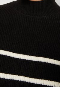 Gros plan sur un pull en tricot côtelé noir avec un col haut et deux bandes blanches horizontales sur la poitrine.