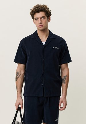Ung mann med krøllete hår iført en mørkeblå kortermet skjorte med knapper og matchende shorts, som holder en svart veske.
