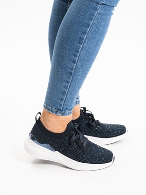 Person wearing black knit sneakers with white soles and blue jeans, standing on a white surface.