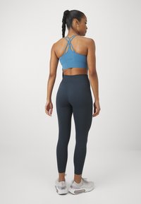 Kék sportmelltartó, keresztezett pántokkal, magas derekú sötét leggingssel párosítva. A modellen világos edzőcipő van, bemutatva a testhez simuló sziluettet.