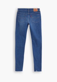 Jean skinny en denim bleu vu de dos avec des poches à double couture, un patch de marque en cuir marron et des ourlets effilochés.