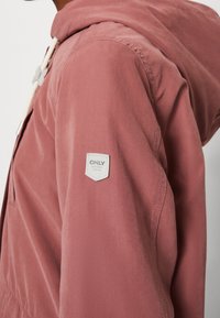 Hellrosa Kapuzenjacke mit strukturierter Oberfläche, versehen mit einem Logopatch am Ärmel und Kordelzügen an der Kapuze.