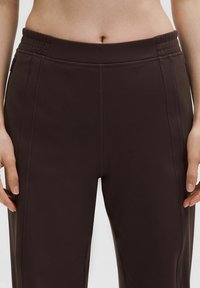 Nahaufnahme einer Person, die hoch taillierte, dunkelbraune Hose mit sichtbaren Nahtdetails und elastischem Bund trägt.