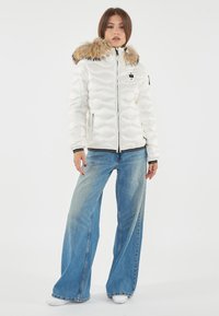 Witte gewatteerde pufferjack met een faux fur-voering in de capuchon, ritssluiting en elastische manchetten, gecombineerd met uitlopende blauwe spijkerbroek en witte sneakers.