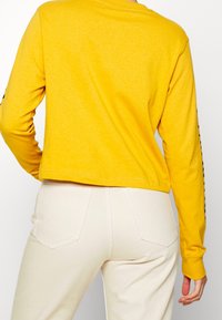 Gul långärmad croppad topp med en mjuk textur, med svarta detaljer längs ärmarna och en avslappnad passform, matchad med krämfärgade jeans.