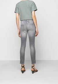 Persona vistiendo jeans skinny gris claro, una blusa verde de manga corta y sandalias de tacón con tiras negras, de pie contra un fondo liso.