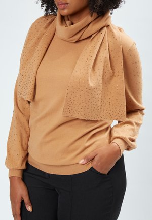 Haut beige à manches longues avec un col drapé et des accents scintillants ; texture douce ; associé à un pantalon noir ; main dans la poche.