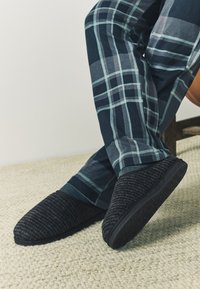 Chaussons noirs texturés avec une semelle noire lisse, associés à un pantalon à carreaux bleu marine et vert, posés sur un tapis de couleur claire.