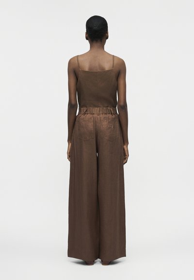 Γυναίκα με σκουρόχρωμο δέρμα και μαλλιά σε κότσο, γυρισμένη προς τα πίσω, φορεί καφέ αμάνικη μπλούζα και φαρδιά παντελόνια σε απλό λευκό φόντο.