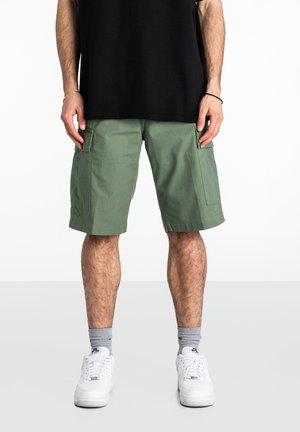 Pantalones cortos cargo en verde oliva, con dos bolsillos laterales, una tela de mezcla de algodón y un corte recto, combinados con zapatillas blancas y calcetines grises.