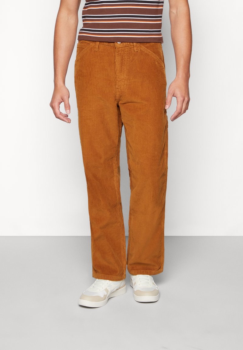 Homme portant un pantalon en velours côtelé marron taille haute avec poches avant, associé à des baskets blanches et un top rayé sans manches.