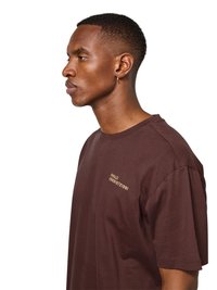 Jonge man met kort haar en een kleine oorring, draagt een bruin T-shirt met gele tekst op de borst, kijkt naar links tegen een witte achtergrond.