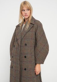 Hnědý kostkovaný kabát s dvojitým zapínáním, velkým klopovým límcem, oversize rukávy a černými knoflíky. Texturovaná látka se vzorem houndstooth.