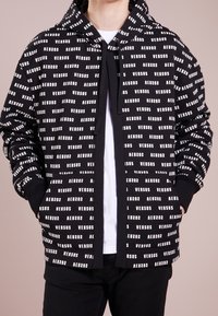 Man som bär en svart zip-up hoodie med vitt "VERSUS" textmönster, vit undertröja och svarta byxor, händerna i hoodiefickorna.