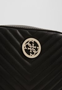 Naert billede af en sort quiltet lædertaske med et guldtonet rundt logo, der viser sammenflettede bogstaver.