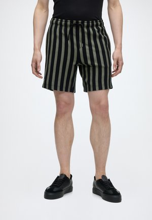 Mand iført sorte og olivengrønne vertikalt stribede shorts, sorte sneakers og en sort top, stående med armene afslappet ned langs siderne.