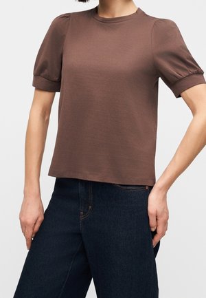 Femme portant un haut marron à manches courtes avec épaules bouffantes associé à un jean taille haute bleu foncé sur un fond uni.
