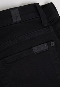 Pantalons en denim noir avec une texture lisse, présentant un design de couture surélevé, une étiquette à la taille et un marquage subtil sur la poche.