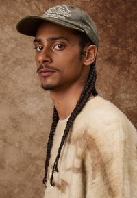 Maslinasto zelena kapa s izvezenim tekstom, nosi se preko bež, teksturirane veste. Model ima pletenice i pozicioniran je protiv neutralne pozadine.