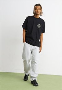 Zwarte katoenen T-shirt met grafisch logo op de borst, gecombineerd met losse witte denim broek en zwarte sneakers. Effen achtergrond.