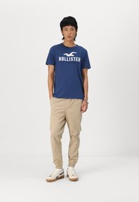 Granatowy t-shirt z białym logo "HOLLISTER", w zestawieniu z beżowymi spodniami dresowymi. Model nosi białe sneakersy z gumową podeszwą. Prosty, codzienny strój.