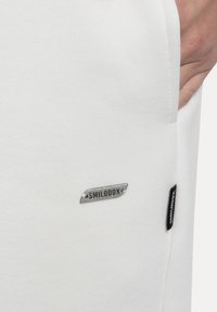 Biała tkanina o gładkiej fakturze posiada srebrną metkę z logo "SMILODOX". Ubranie ma kieszeń z boku.