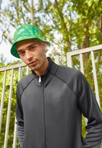 Giacca atletica nera con zip e cuciture grigie, caratterizzata da un logo discreto. Cappello a secchiello con motivo verde. Sfondo naturale con vegetazione.