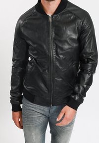 Man som bär en svart läderbomberjacka med dragkedja och ribbade manschetter, i kombination med ljusblå slitna jeans.