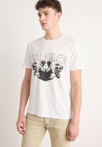 Hvit bomulls T-skjorte med et svart grafisk motiv av to løvehoder og ordet "HUGO" ovenfor. Klassisk passform med rund hals.