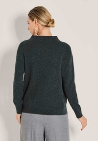 Frau mit blonden Haaren im Dutt trägt einen dunkelgrauen Pullover und hellgraue Hosen, steht mit dem Rücken zu einer schlichten hellen Wand.