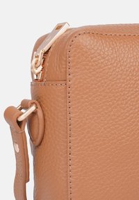 Zbliżenie na róg torebki z jasnobrązowej skóry, ukazujące teksturę ziarna, złoty suwak zamka oraz przymocowany pasek ze złotymi okuciami.