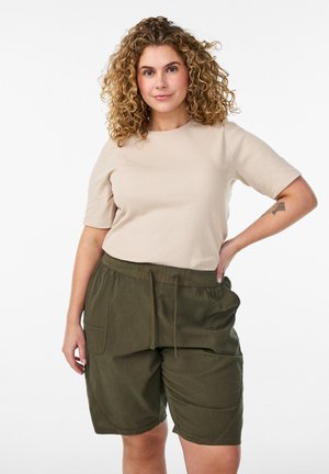 Krullendharige vrouw met een beige T-shirt met korte mouwen en olijfgroene shorts met trekkoord, staand met één hand op haar heup tegen een witte achtergrond.