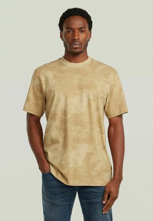 Uomo con capelli corti e ricci, che indossa una camicia beige a maniche corte con texture e jeans blu, in piedi con una mano in tasca su sfondo neutro.