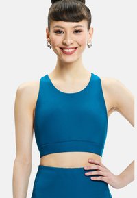 Young woman smiling with hand on hip, wearing blue sleeveless crop top and matching high-waisted bottoms against plain background.