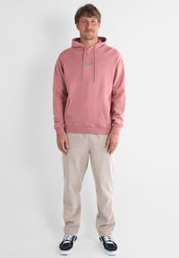 Felpa rosa con cappuccio, cordini, tasca frontale e dettaglio logo; abbinata a pantaloni in corduroy beige e sneakers nere. Outfit semplice e casual.