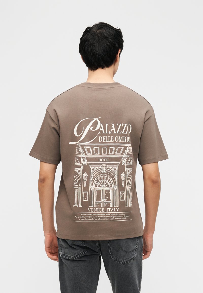 Άνδρας που φοράει καφέ μπλουζάκι με λευκό σκίτσο αρχιτεκτονικού ξενοδοχείου και κείμενο «Palazzo Delle Ombre, Βενετία, Ιταλία» στην πλάτη.