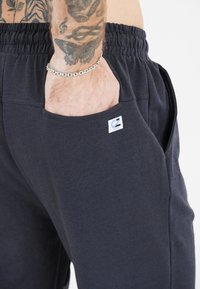 Tumma harmaat puuvillajoggerit, joissa on joustava vyötärö, sivutaskut ja pieni brändilogo-patch. Sileä pinta ja rento muotoilu.