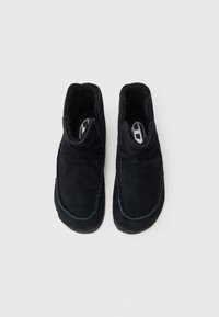 Zapatos negros de ante tipo slip-on con un interior forrado suave, detalles de costura sutiles y una forma de punta redonda. Sin hardware ni patrones visibles.