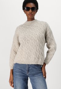Kabelstrikket genser i lys grå, med høy hals og ribbestrikket fald. Brukt med blå jeans og kombinert med svarte solbriller.