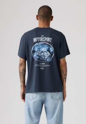 Mand med tatoveringer iført marineblå Levi's Motorsport T-shirt med motorcykelgrafik og lyseblåt jeans, vendt væk fra ensfarvet baggrund.
