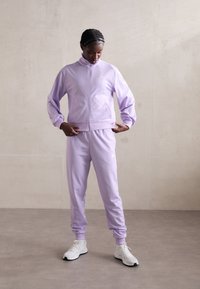 Λιλά σετ φόρμας με τζάκετ που κλείνει με φερμουάρ και ταιριαστά παντελόνια. Φτιαγμένο από λείο ύφασμα, με ριμπ μανσέτες και λευκές λεπτομέρειες με λογότυπο.