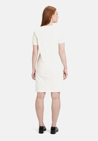 Weißes Kleid mit kurzen Ärmeln, strukturiertem Stoff, knielang, mit einem einfachen, klaren Design und gerader Silhouette. Sichtbare schwarze Schuhe.