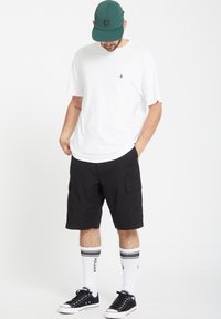 Weißes T-Shirt mit kurzen Ärmeln, schwarze Cargo-Shorts, grüne Kappe, schwarze Sneakers und weiße Socken mit schwarzen Streifen. Einfacher, lässiger Streetwear-Stil.