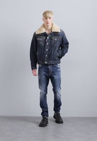 Young man standing, wearing dark denim jacket with beige shearling collar, faded blue jeans, and black lace-up shoes against plain gray wall.