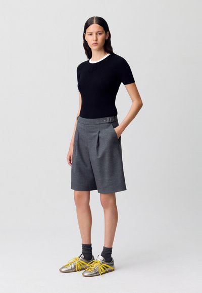 Top negro ajustado con cuello blanco, pantalones cortos gris a medida con pliegues, y zapatillas metálicas con cordones amarillos y calcetines grises.