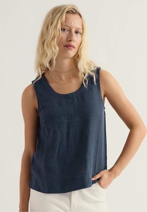 Heledate juustega blond naine, kes kannab tumenäost varrukateta topi ja valgeid pükse, poseerib ühe käega pihikusse tõstetuna neutraalse taustaga.