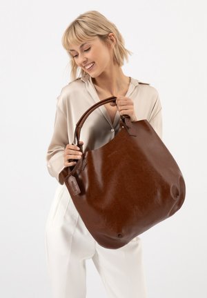 Lachende vrouw met kort blond haar houdt een grote bruine leren draagtas vast, draagt een beige blouse en witte broek tegen een effen witte achtergrond.