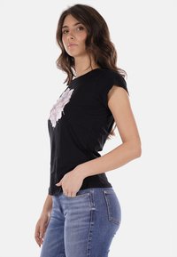 T-shirt nera a maniche corte con una grafica floreale testurizzata in rosa e bianco sul davanti, abbinata a jeans in denim blu aderenti.