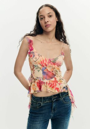 Jeune femme aux longs cheveux bouclés portant un haut à volants sans manches à fleurs et un jean bleu, debout les mains derrière le dos.